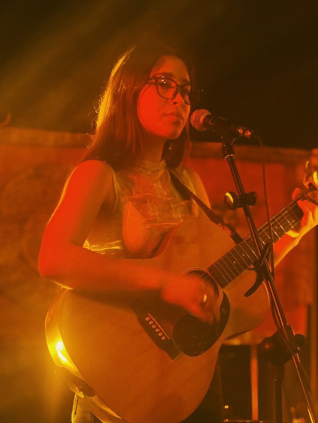 A woman with glasses playing an acoustic guitar on stage, illuminated by warm yellow and orange lighting.