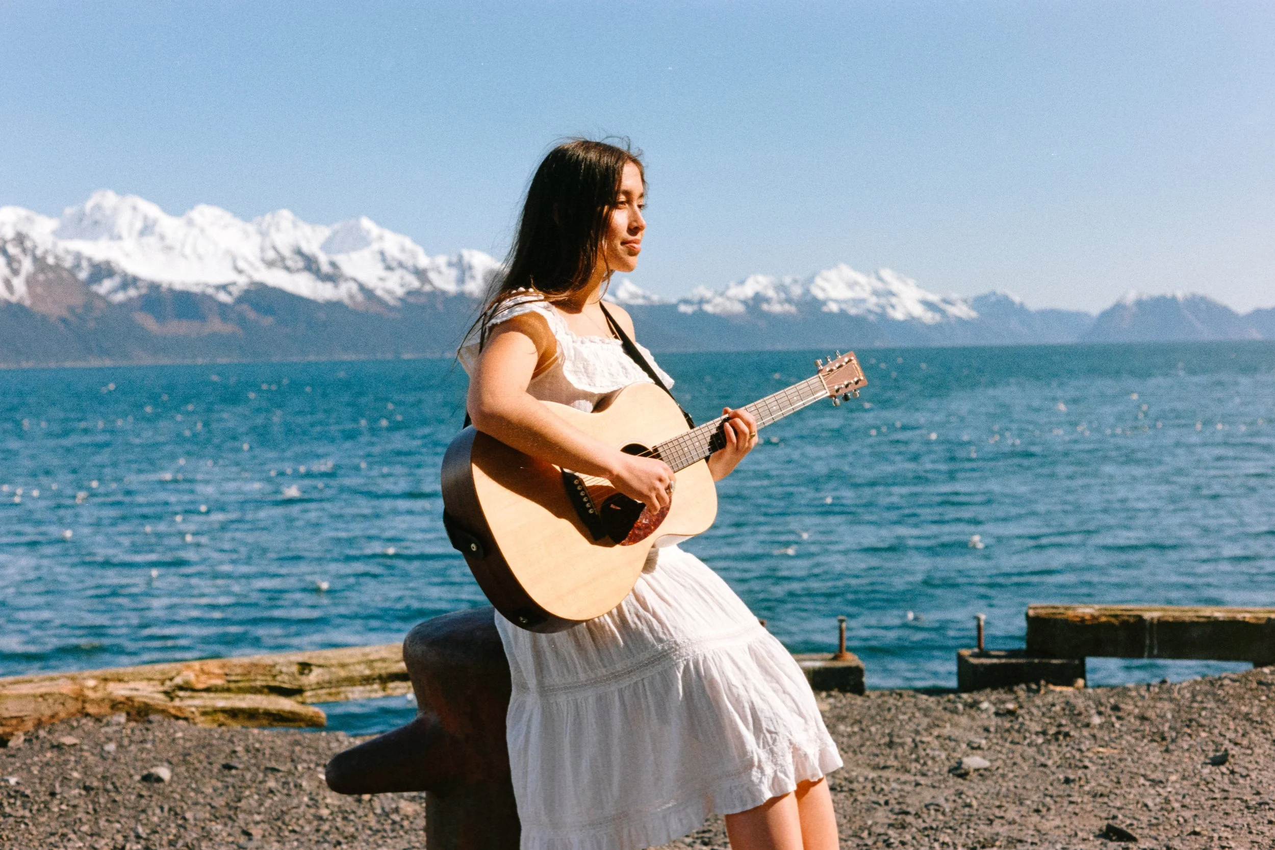 Young woman with long dark hair playing an acoustic guitar near a body of water with snow-capped mountains in the background.