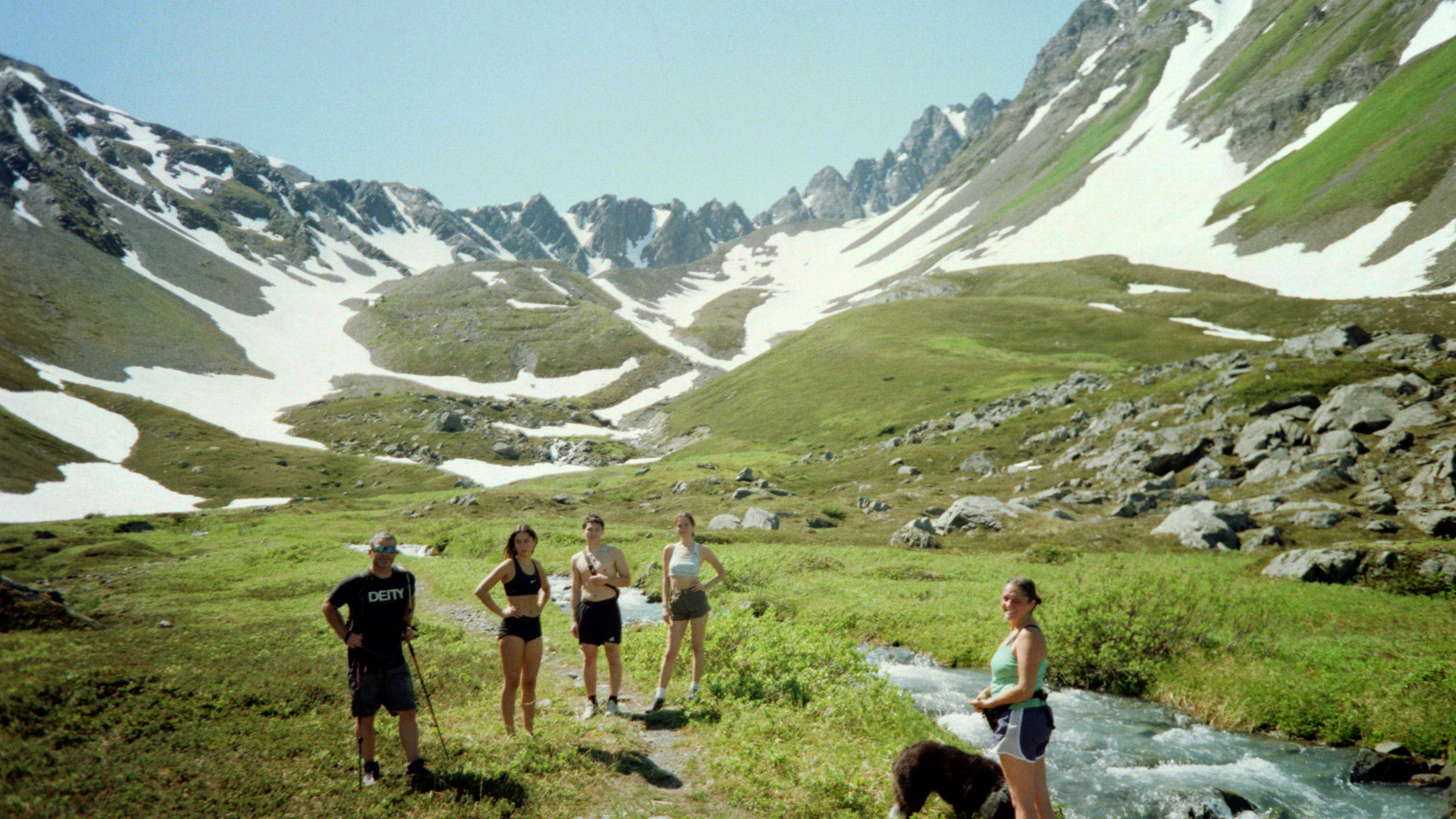 Four young adults standing near a mountain stream in a lush green valley with snow-capped mountains in the background.