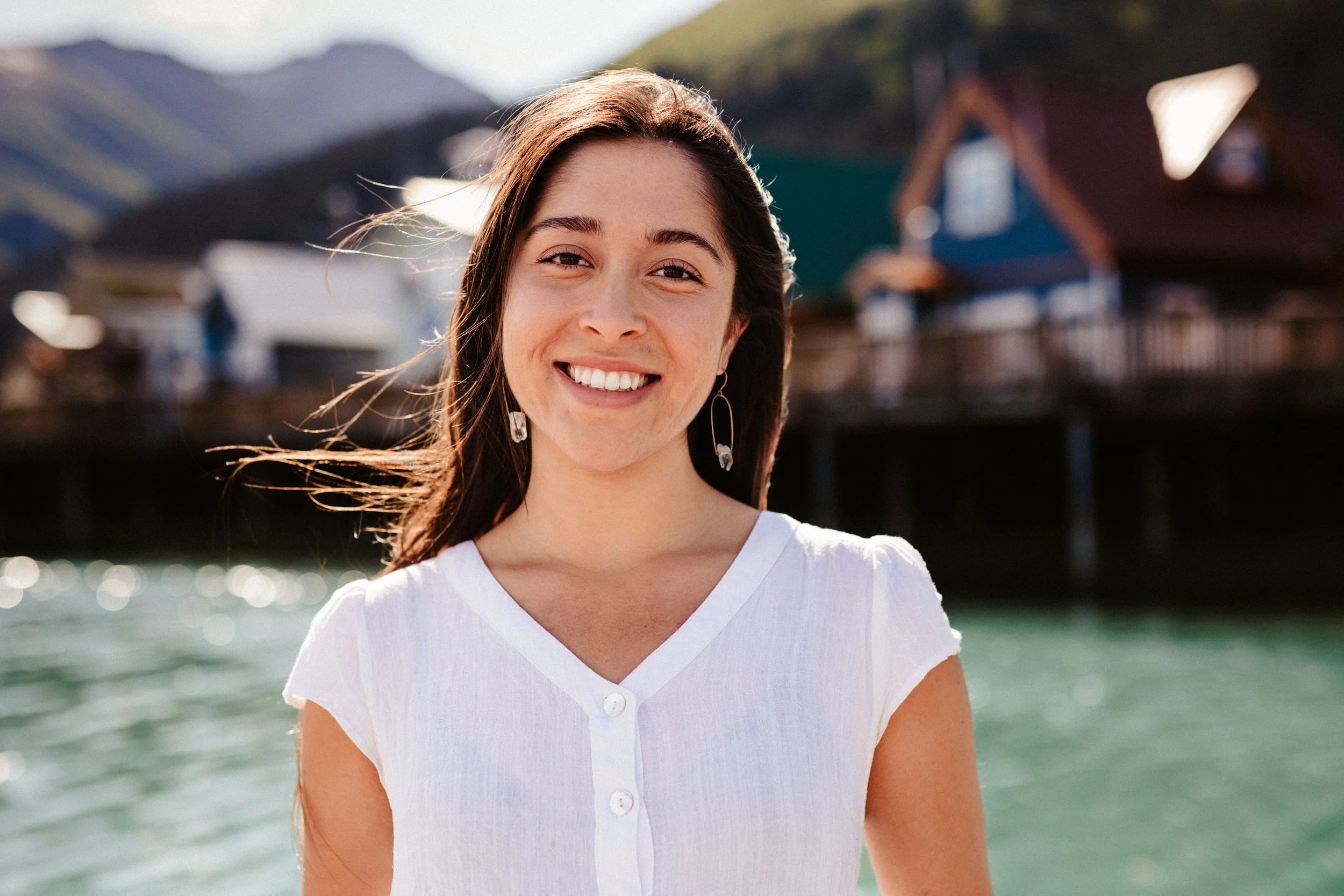 A smiling woman with long brown hair standing outdoors near water, with mountains and buildings in the background during daytime.
