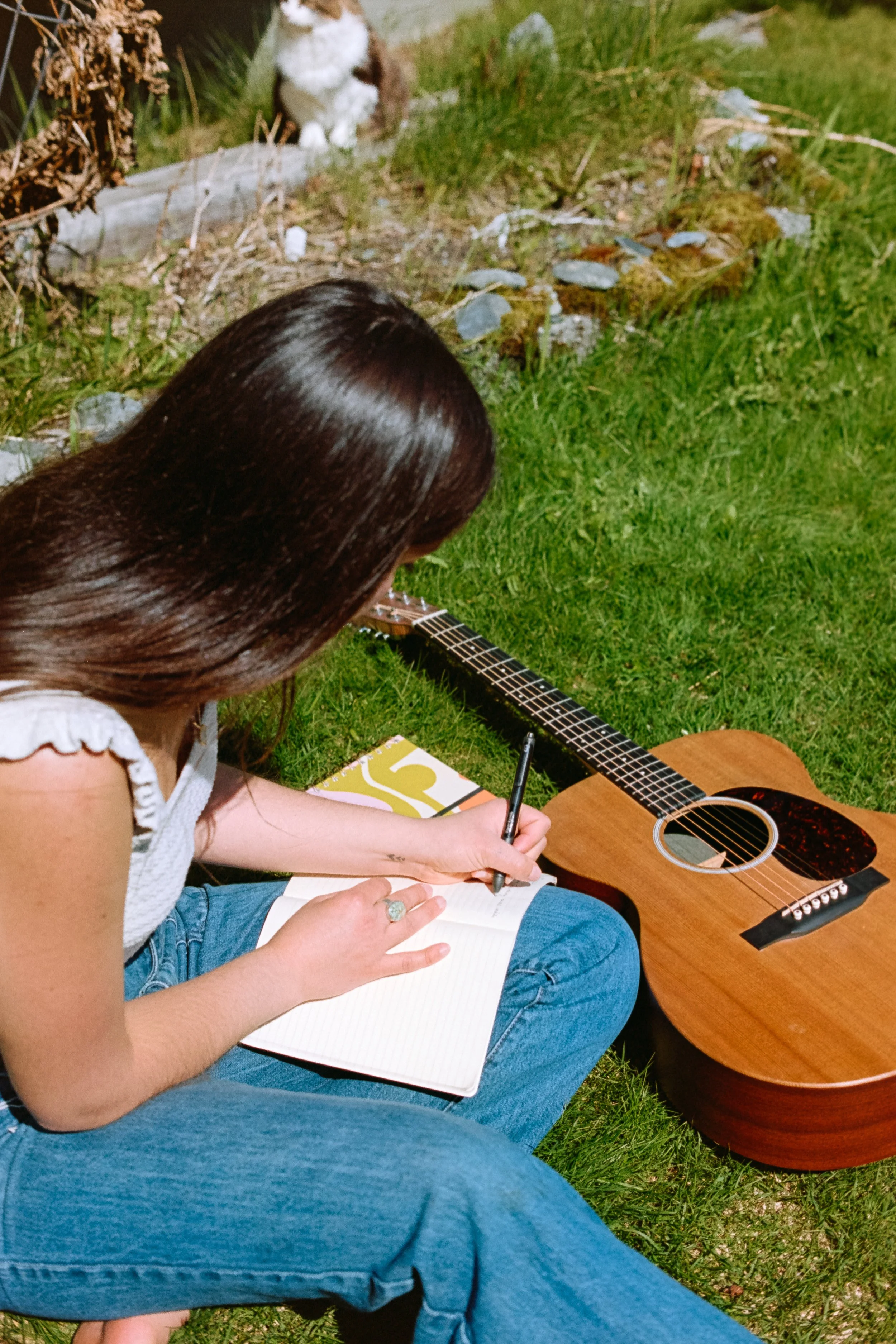 A young woman with long brown hair, wearing a white sleeveless top and blue jeans, is sitting on grass outdoors with a notebook on her lap, writing with a pen. Next to her, there is an acoustic guitar on the grass. In the background, here is a garden with greenery and rocks.