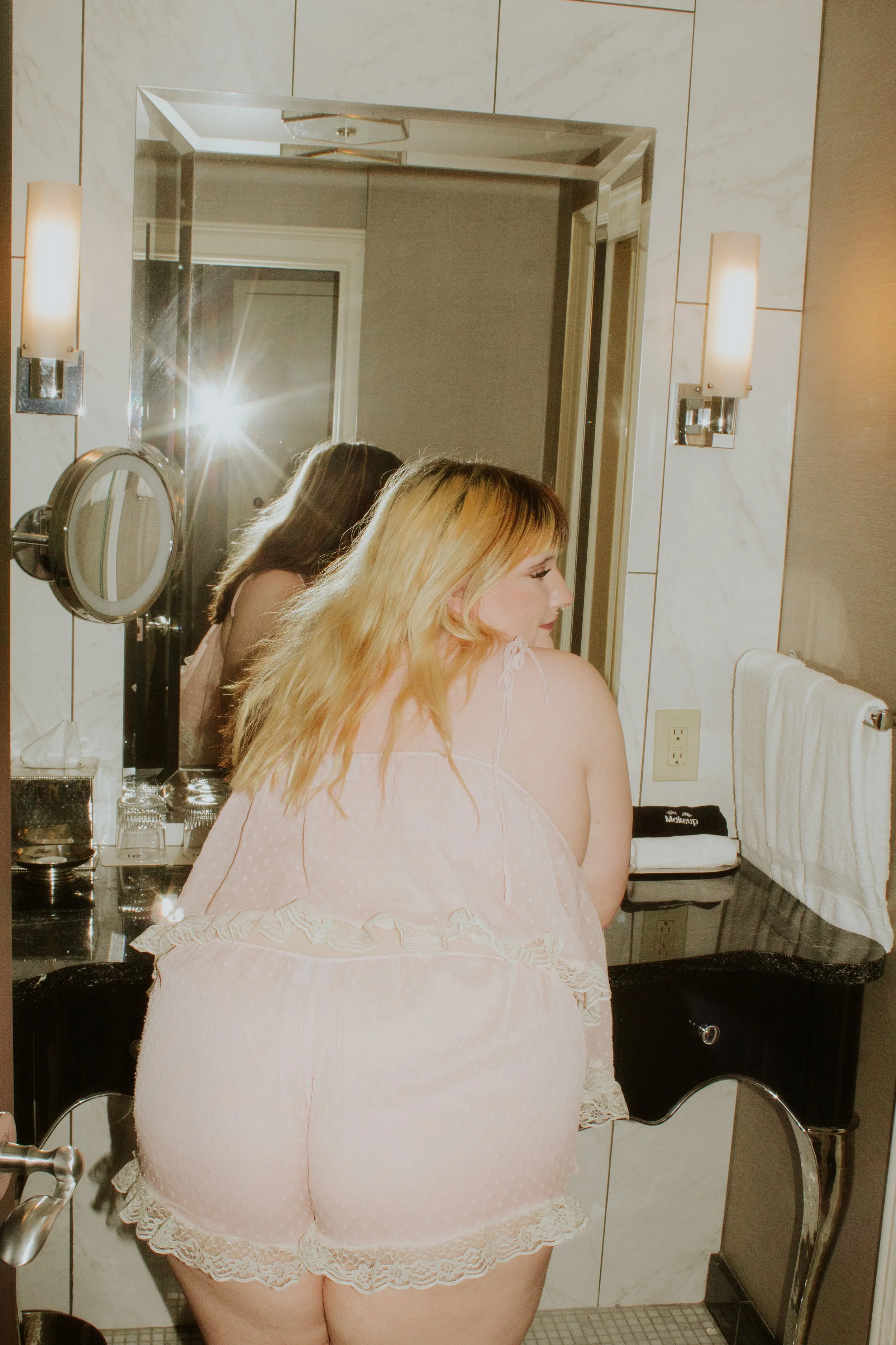 A woman with blonde hair looking in a bathroom mirror, wearing pink lace pajamas, with a bathroom sink and wall-mounted lights visible.