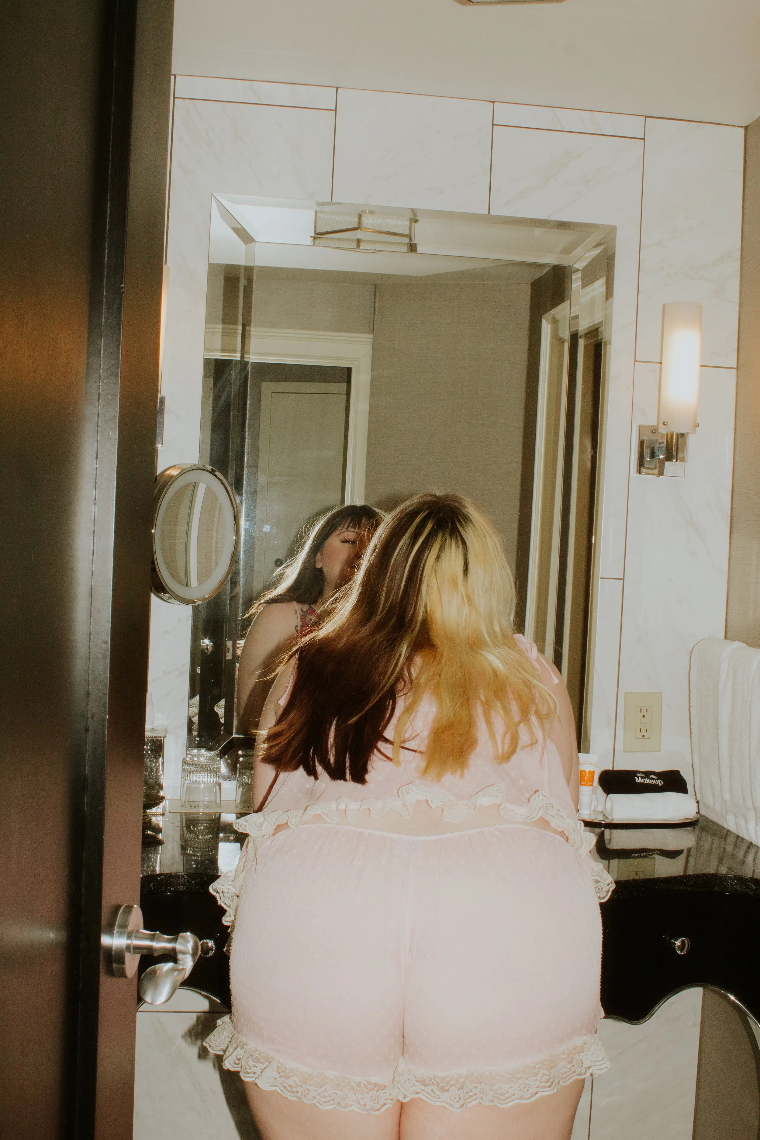 A woman with long, light brown and blonde hair sitting on a white, lace-trimmed plush chair in front of a large mirror in a bathroom. The bathroom has white marble walls with gold accents, a rectangular mirror, and a wall-mounted light fixture. On th