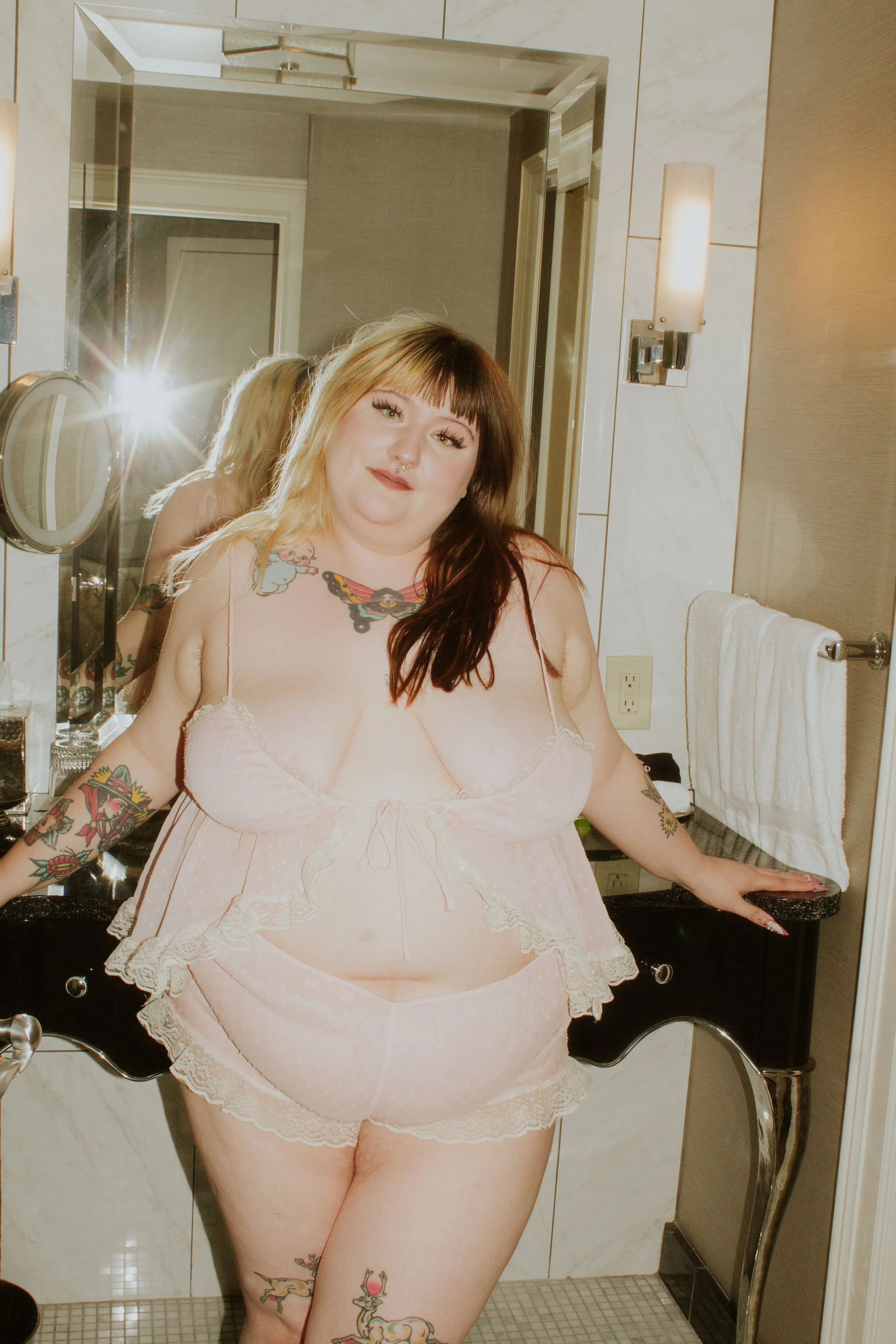 A woman in pink lingerie with tattoos stands in front of a bathroom mirror, smiling softly, with a towel hanging on a rack and bathroom fixtures visible in the background.