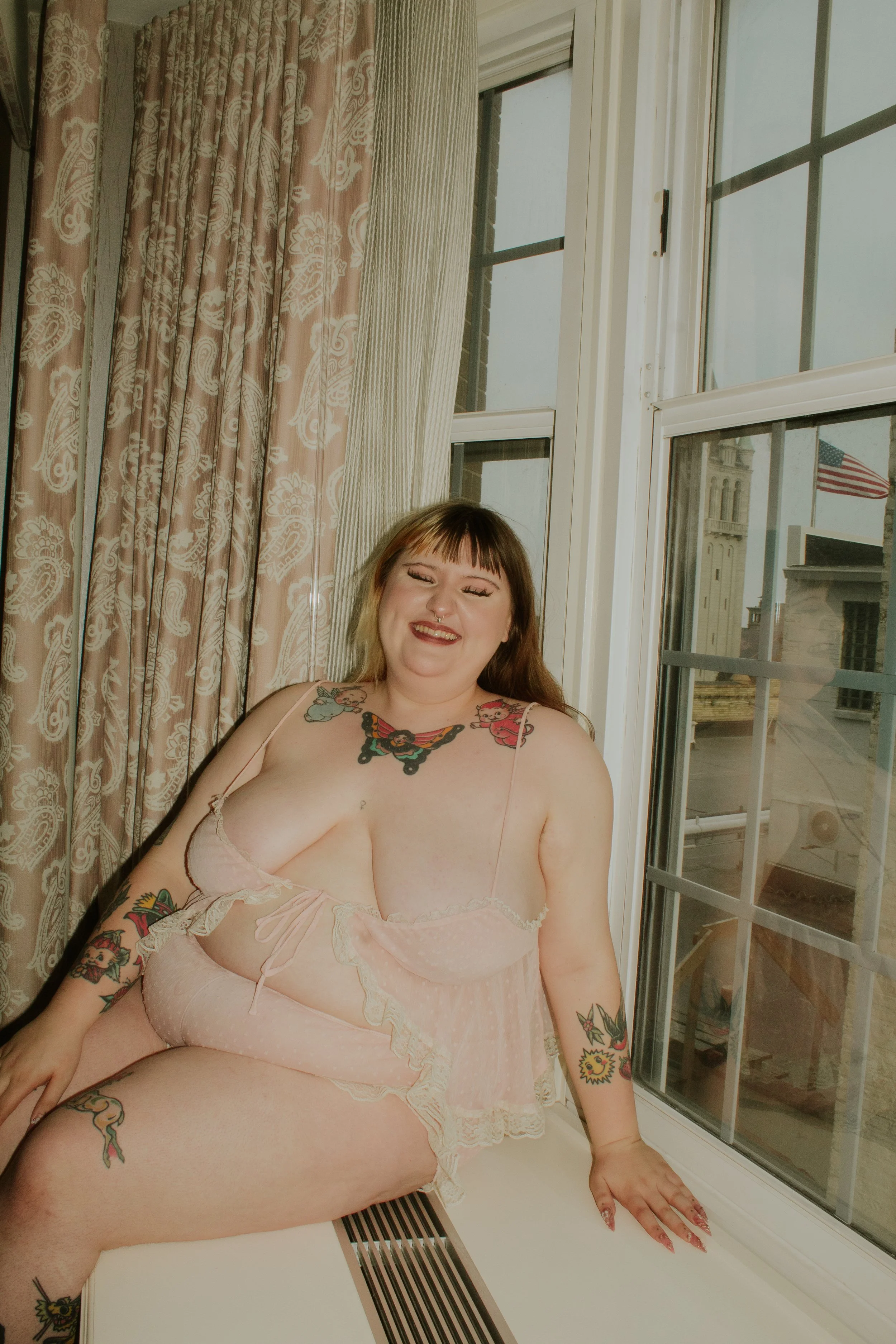A woman with tattoos smiling and sitting by a large window, wearing light pink lingerie, in a room with beige curtains and an American flag visible outside.