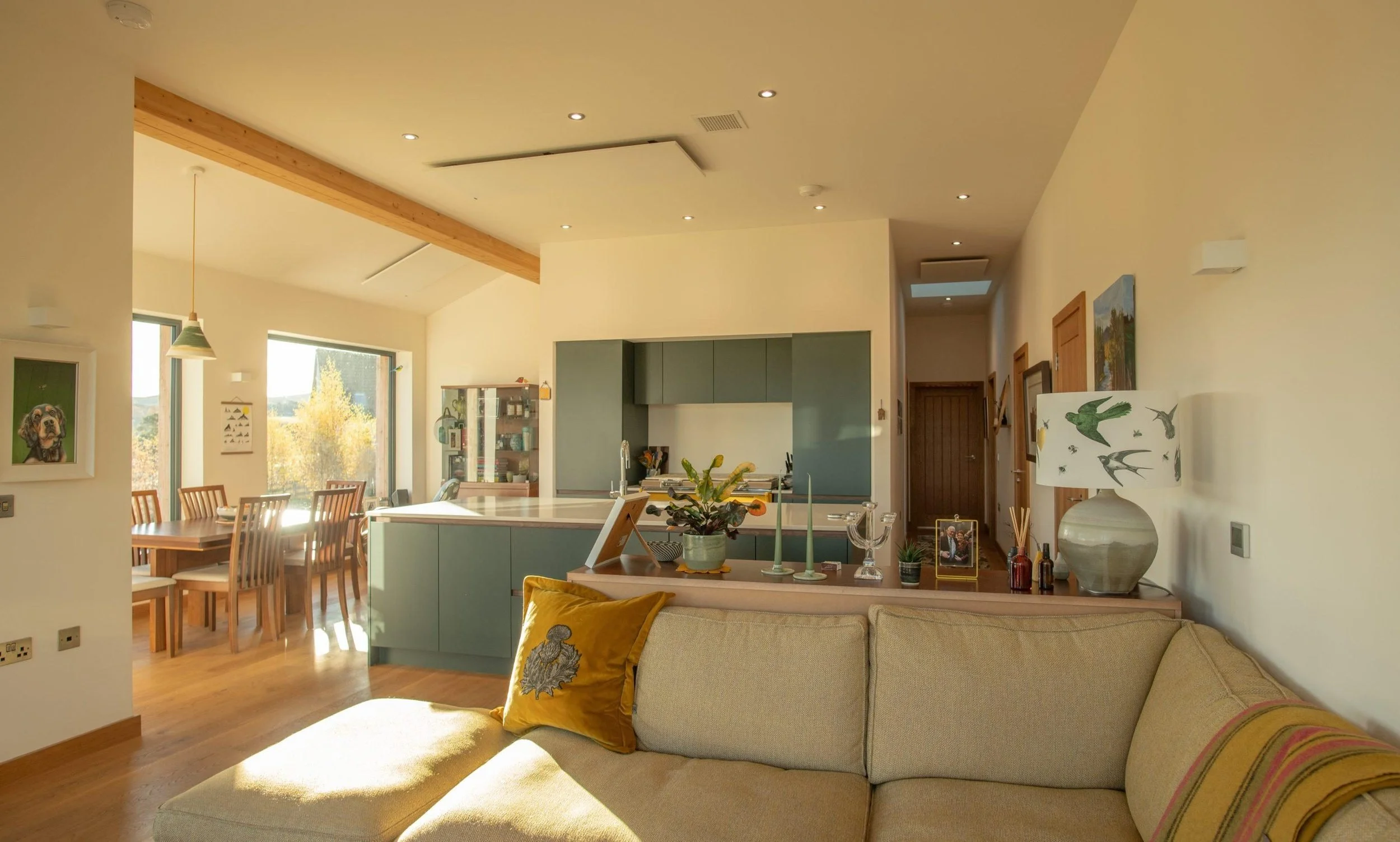 An open plan living room and kitchen area with a beige sofa, colourful pillows, a breakfast table with chairs, and a green kitchen island. Bright sunlight streams through large windows in this MAKAR timber home design.