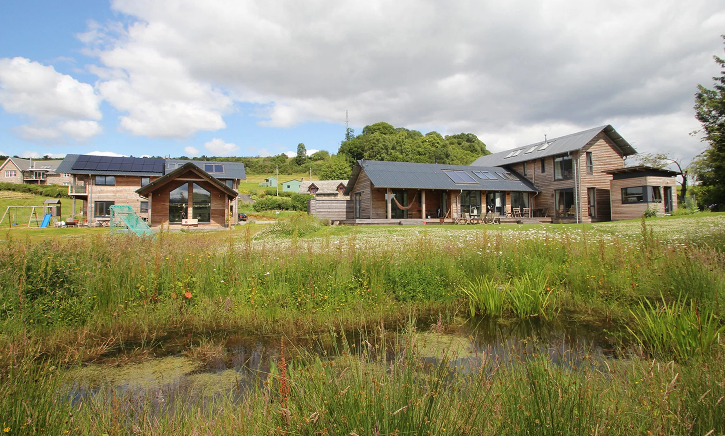 A rural landscape with two MAKAR timber houses, a small pond, and a grassy area with wildflowers under a partly cloudy sky.