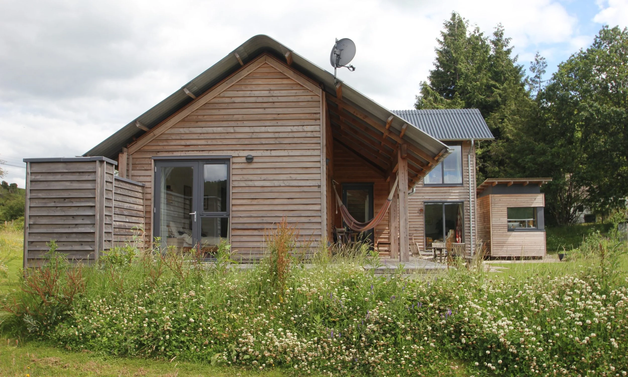 Wooden house with large glass doors on a grassy area with wildflowers, surrounded by trees and a partly cloudy sky.