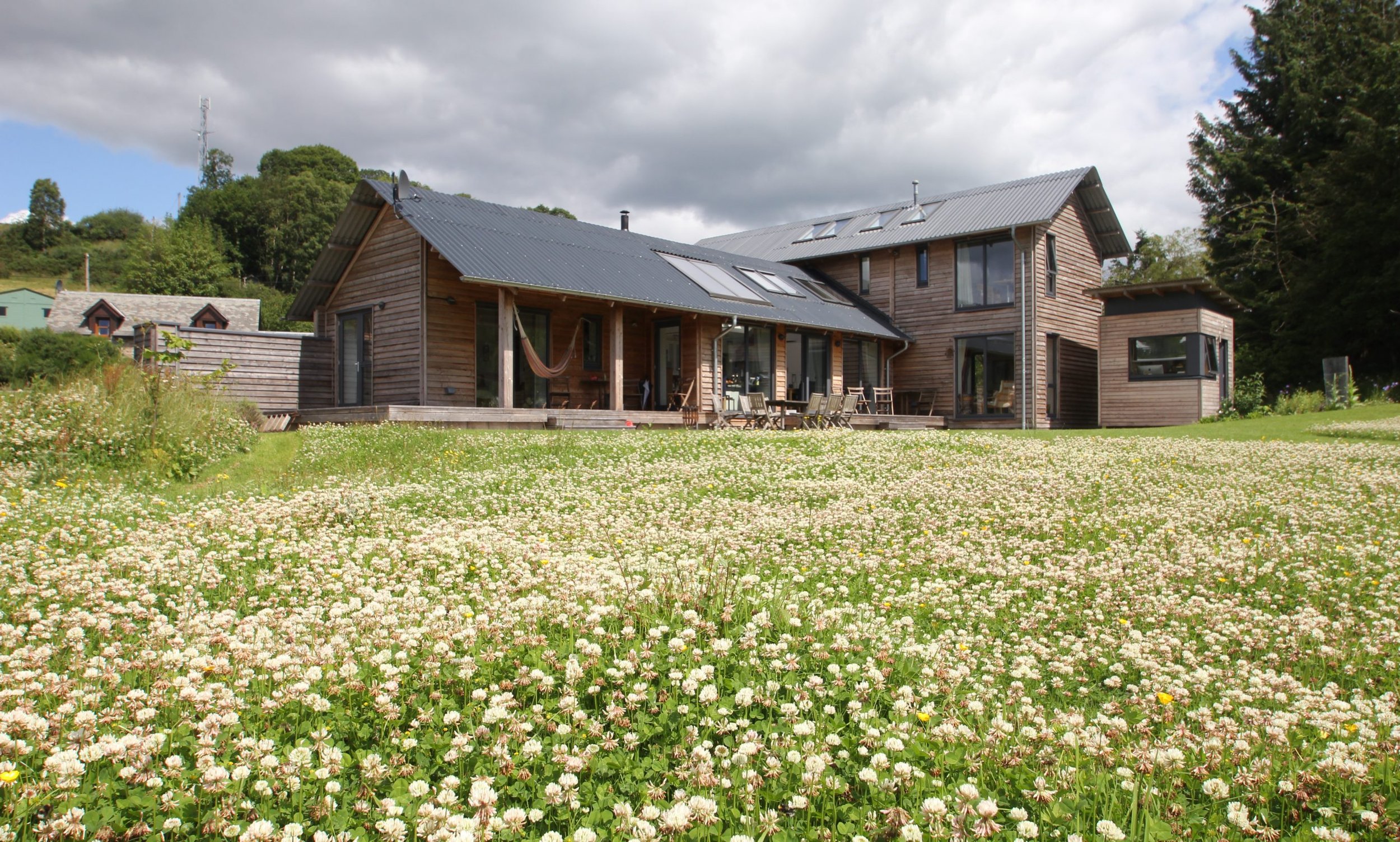 A modern and large MAKAR timber home with a gray metal roof, surrounded by a field of blooming white flowers and greenery, under a cloudy sky.