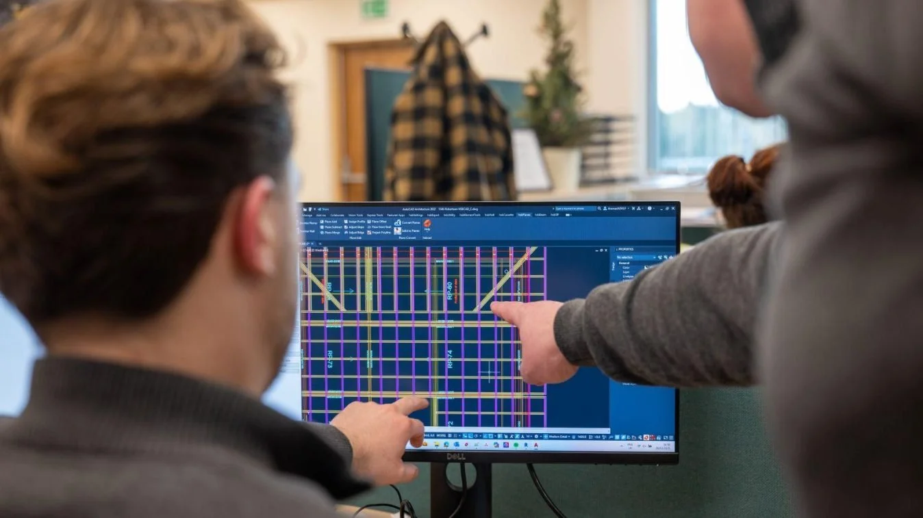 Two people working on a computer with a technical diagram on the screen, for a home-build, in a bright office.