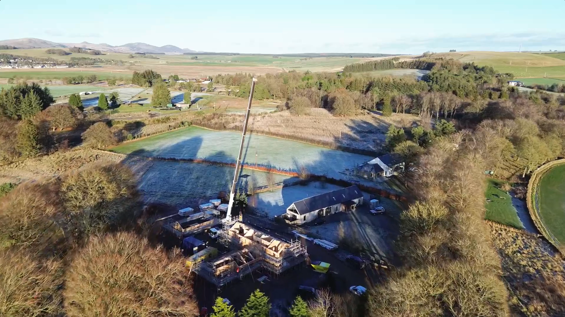 Aerial view of a MAKAR construction site with a crane, surrounded by trees, residential buildings, and farmland with dry and green fields. Mountains can be seen in the background.