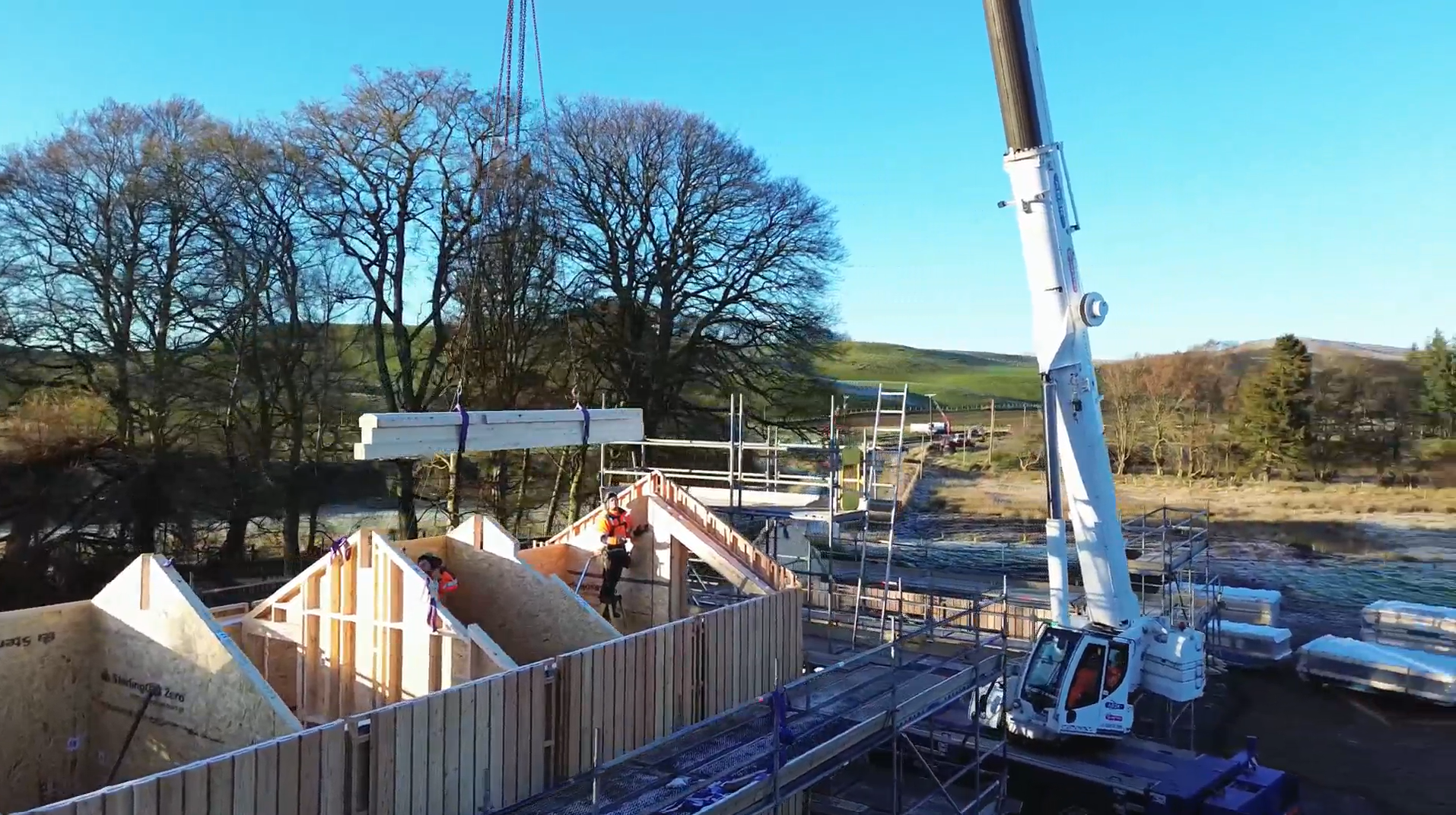 MAKAR construction site with workers in safety gear building wooden structures, crane lifting materials, and trees in the background under a clear blue sky.