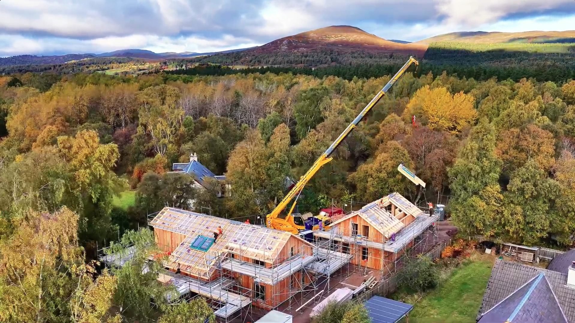 An aerial view of a MAKAR home under construction surrounded by trees in autumn with a crane on site, mountains in the background, and partly cloudy sky.