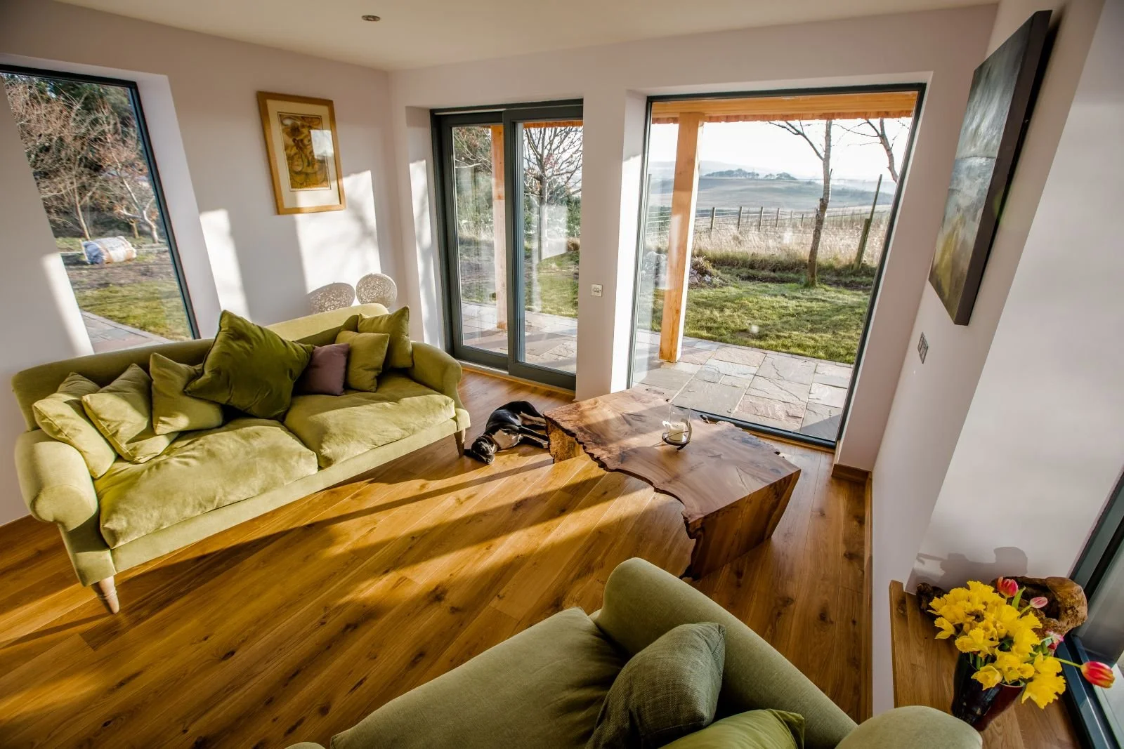 Living room with green sofas, wooden floor, large windows, and a view of the garden and countryside outside. There is a rustic wooden coffee table and a vase with yellow flowers.