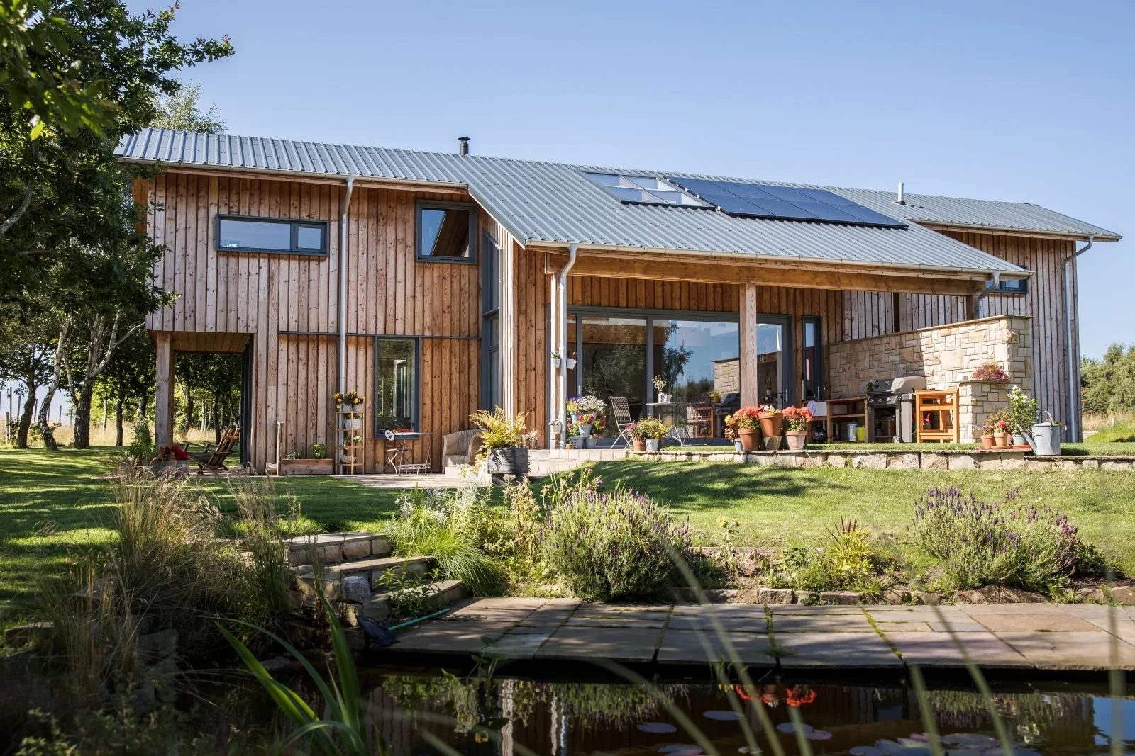 A modern timber MAKAR home with a sloped metal roof, solar panels, large glass sliding door, and outdoor patio with plants and furniture. The house is surrounded by lush green grass and trees, with a small pond in the foreground.