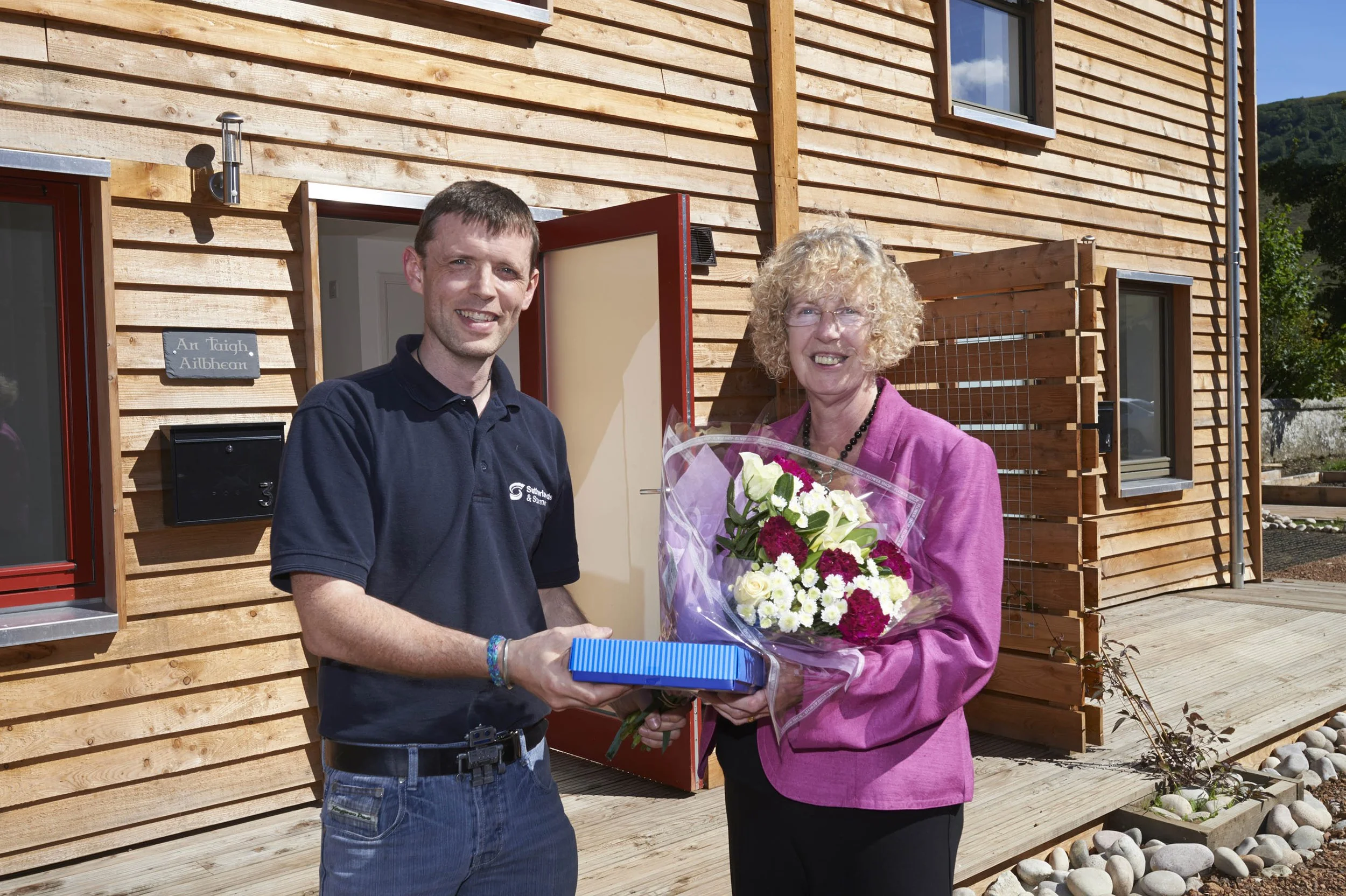 A man and a woman standing outside timber MAKAR affordable homes. The man handing the woman a wrapped gift and the woman holding a bouquet of white and red flowers, both smiling.