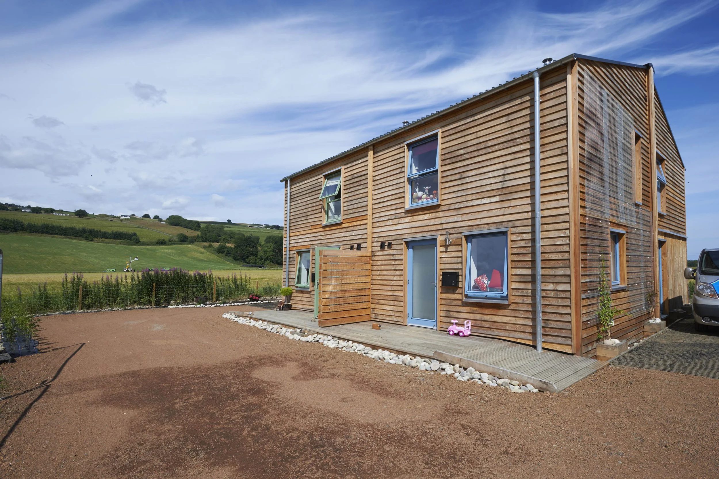 Two semi-detached timber affordable four-bedroom MAKAR homes. With multiple windows with various decorations, a small outdoor patio, and a pink toy car parked nearby, set against a rural landscape with green rolling hills under a partly cloudy sky.