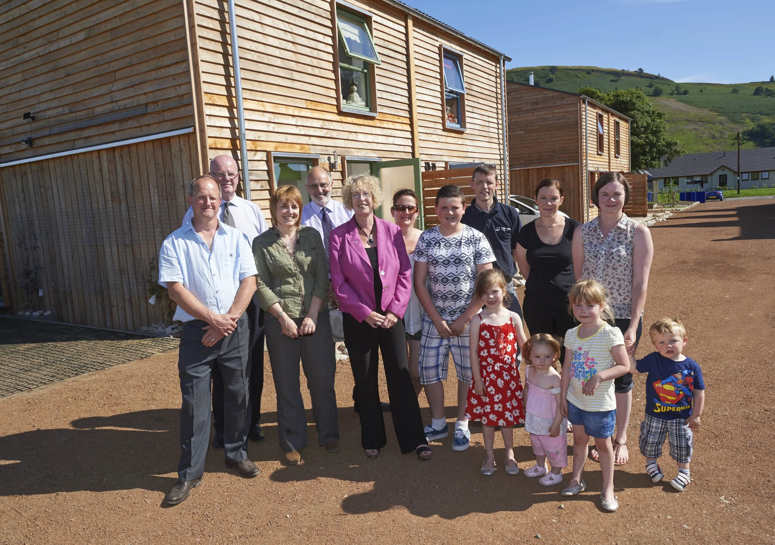 A group of adults and children standing outside in front of newly built wooden houses on a sunny day.