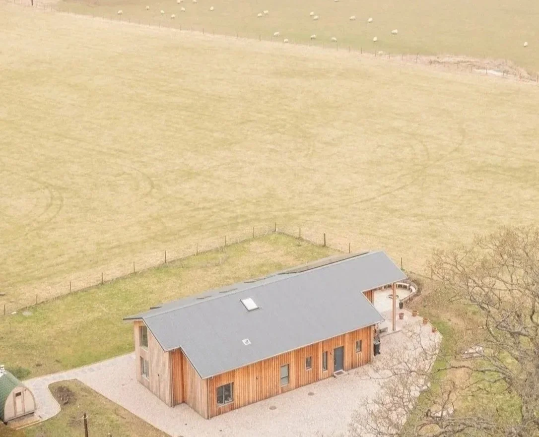 A wooden house with a gray roof is surrounded by a grassy yard and trees, with open fields in the background and a small shed nearby.