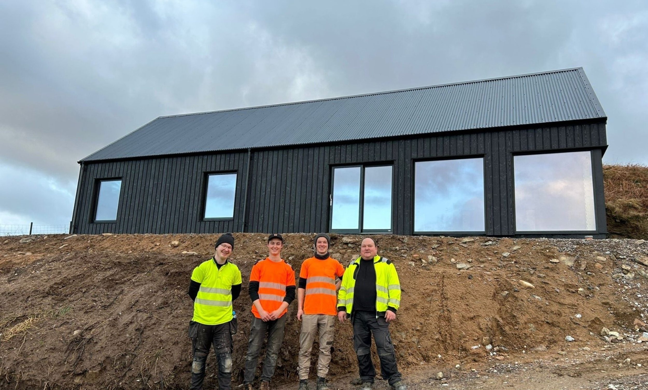 Four construction workers stand in front of a newly assembled modern black MAKAR home, with cloudy sky overhead.