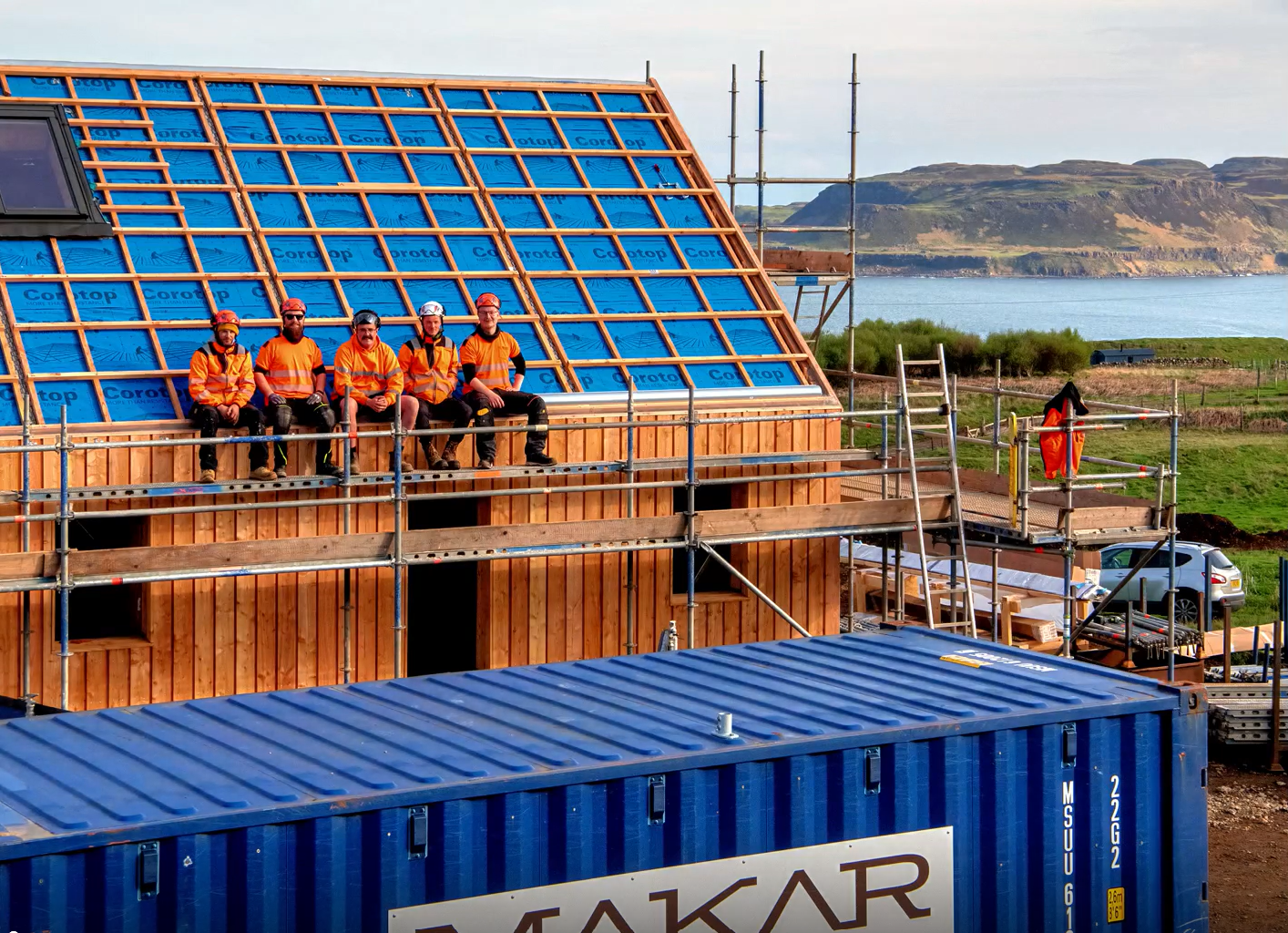 Five construction workers in orange safety vests and helmets sit on scaffolding on a building under construction. The building has a wooden frame and blue insulation panels. In the background, there is a scenic view of a body of water and distant cli