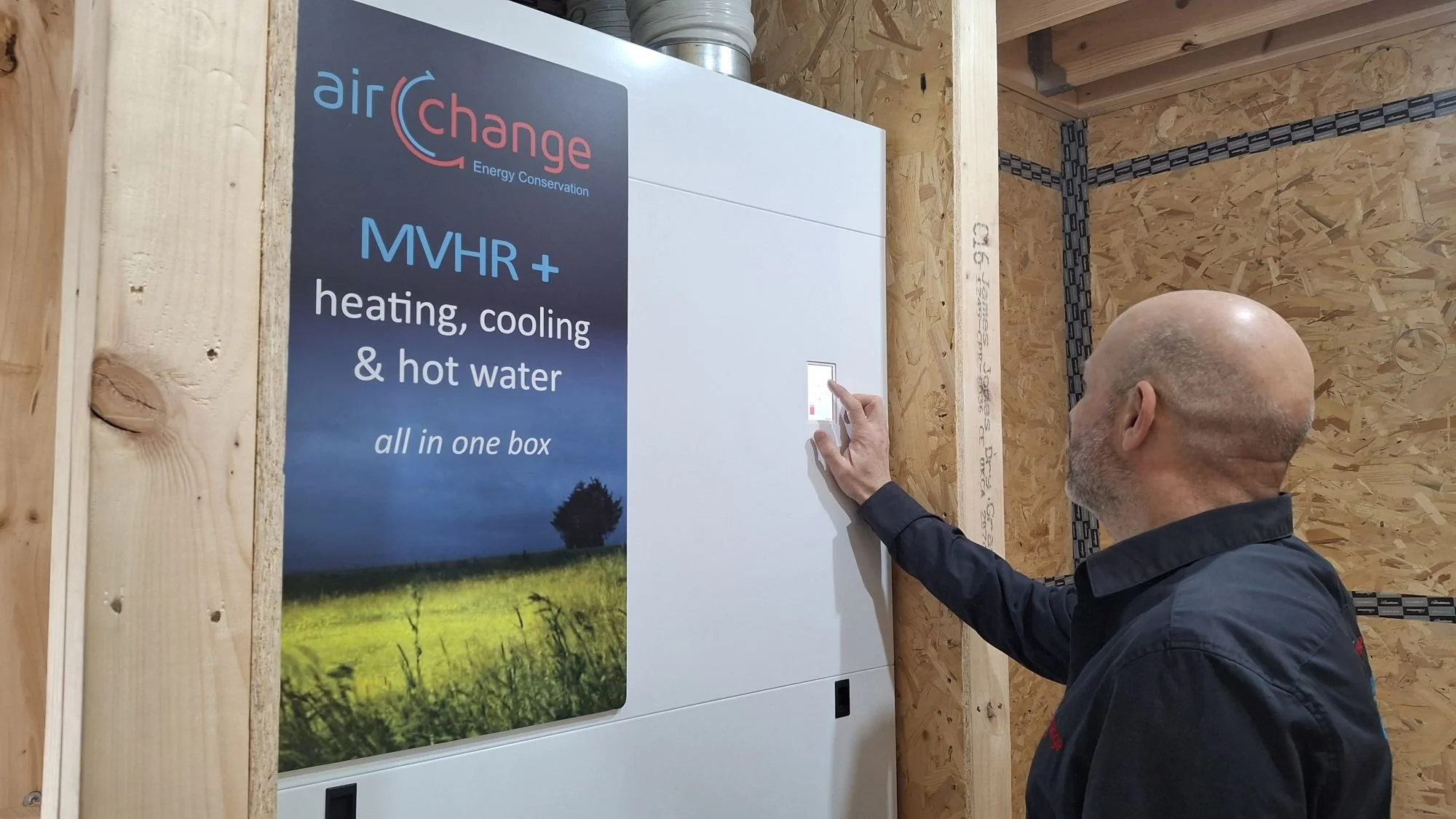 Man adjusting a control panel for a heating, cooling, and hot water system, including MVHR, in a wooden room, with a sign describing the system's functions.