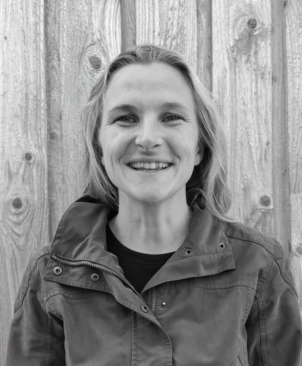 A woman with long, wavy hair smiling at the camera, standing in front of a wooden fence, wearing a dark jacket.