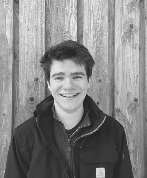 A young man smiling, wearing a jacket, standing in front of a wooden fence.
