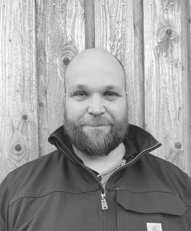 A smiling man with a beard and shaved head wearing a jacket, standing in front of a wooden fence.