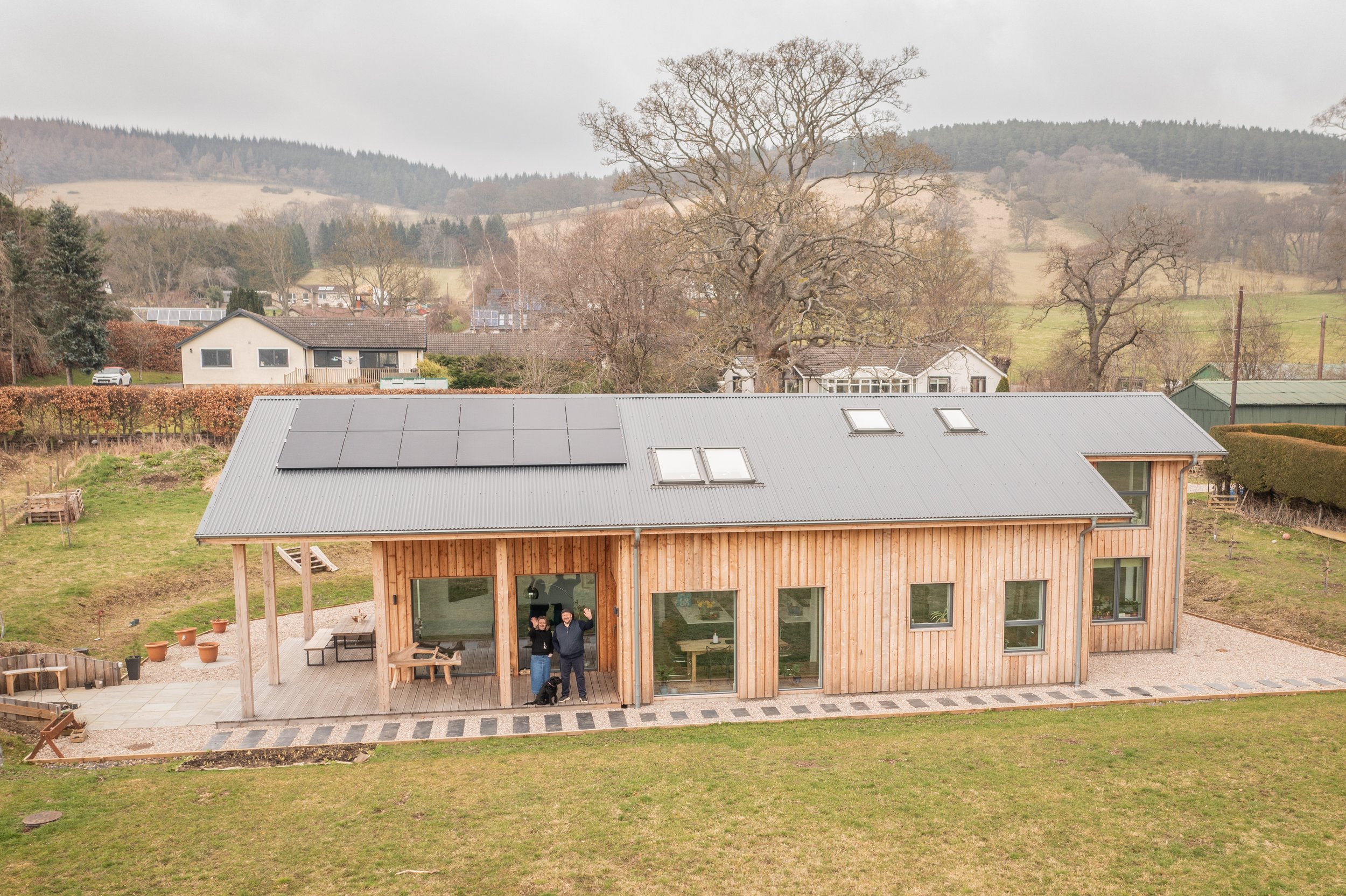 A modern timber MAKAR home with solar panels on the roof and large windows, two people standing on the front porch waving, surrounded by a grassy yard and neighbouring houses with rolling hills in the background.