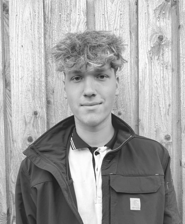 Young man with curly hair, smiling slightly, wearing a jacket over a collared shirt, standing in front of a wooden fence.
