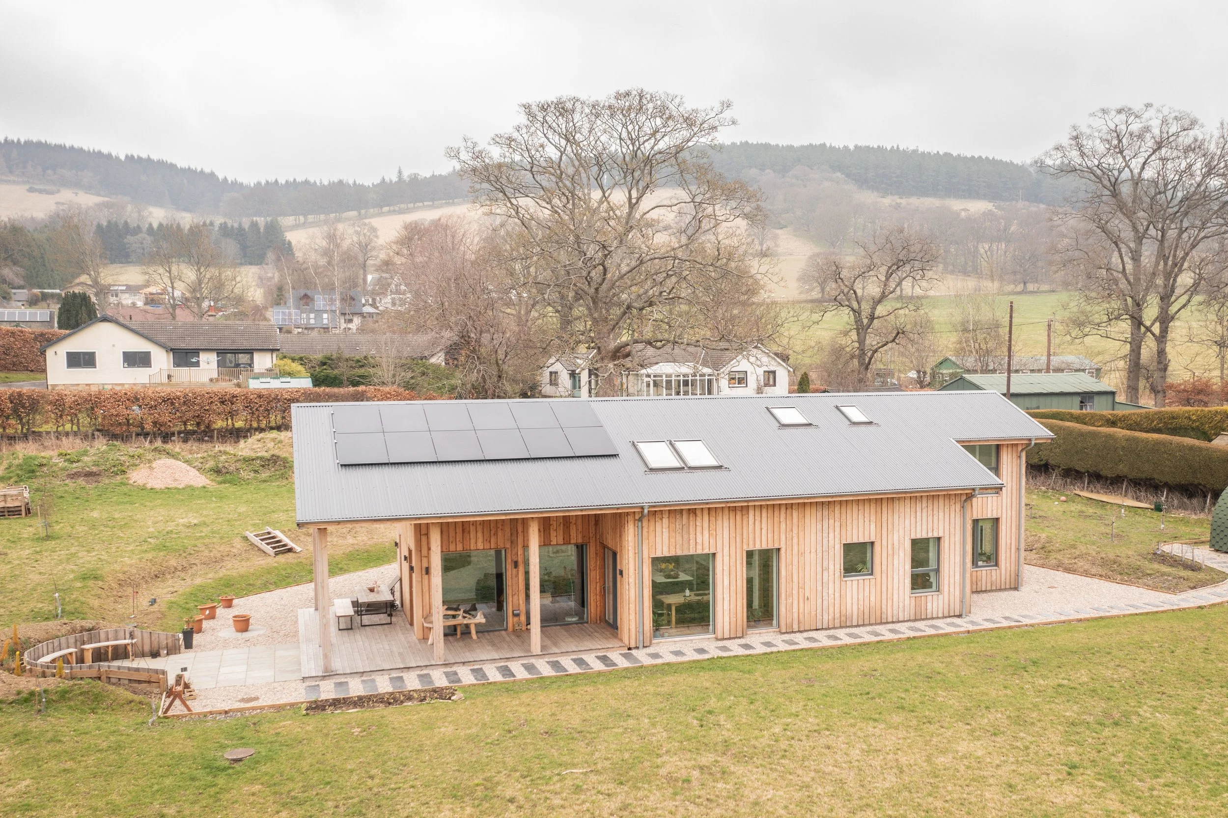 A modern timber MAKAR house with a metal roof and large windows, surrounded by a grassy yard with some trees and neighboring houses in the background, overcast sky.