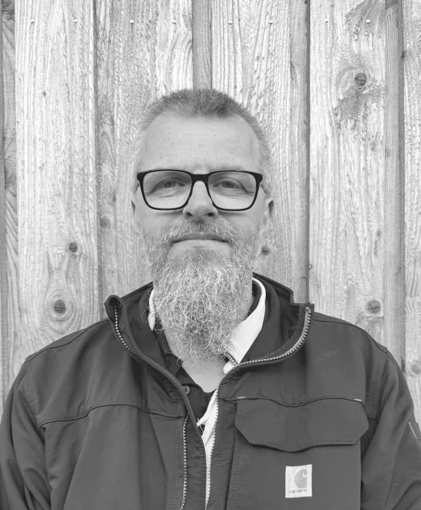 A middle-aged man with glasses and a beard stands in front of a wooden wall, wearing a black jacket.