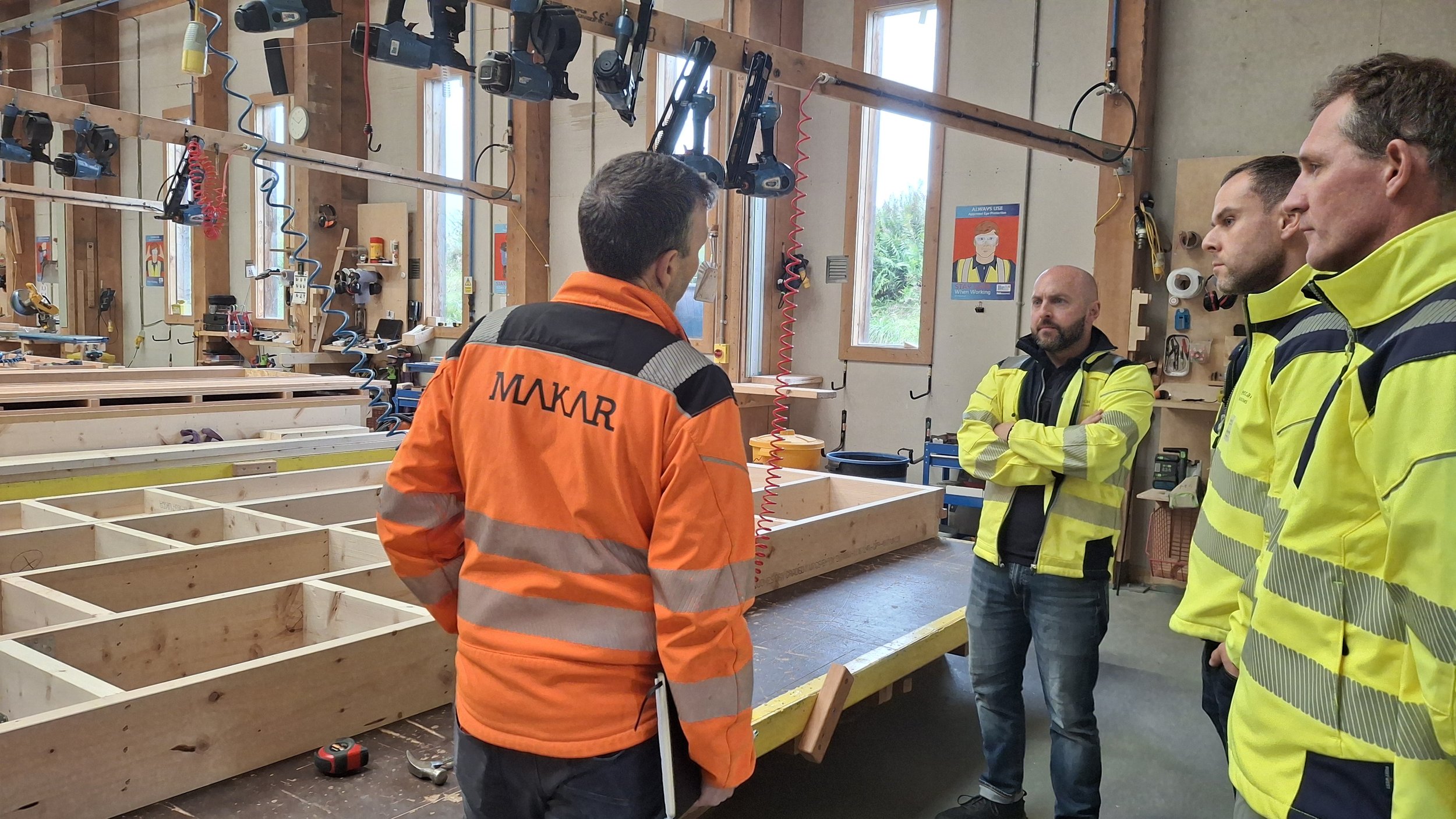 Four workers in safety jackets inside MAKAR's manufacturing workshop. One worker in an orange jacket gives a tour to three people in yellow jackets. Partially assembled closed panels for home construction are in the background.