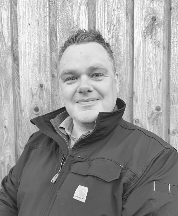 A man with short hair smiling, wearing a jacket over a collared shirt, standing in front of a wooden fence.