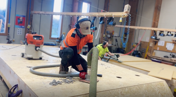 Two construction workers in an indoor workshop, one in an orange safety vest and gray hat using an insulation hose on the closed panel, with another worker in a yellow shirt working in the background.