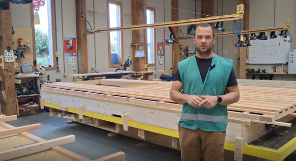 A man standing in a woodworking workshop with lumber and tools, wearing a teal safety vest.