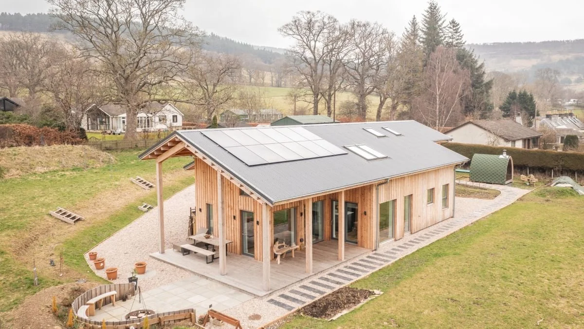 A modern MAKAR sustainable home with a slanted roof with solar panels, large windows, and a covered patio area, situated in a rural area with trees and other houses in the background.