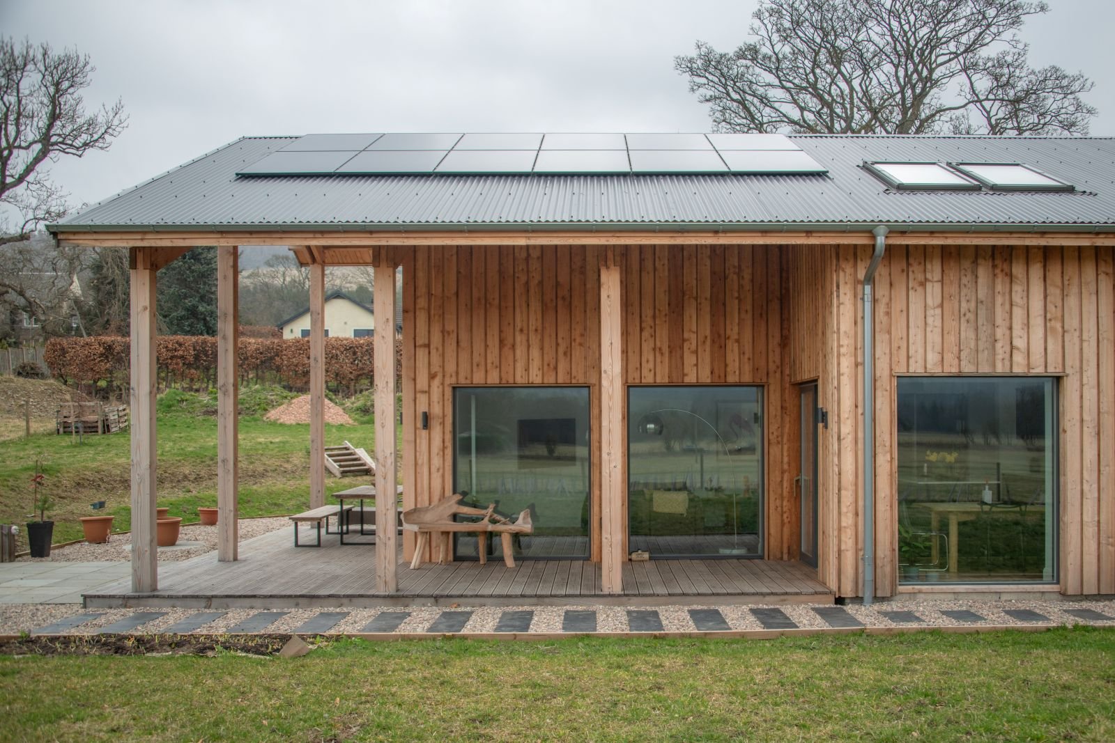 View of a modern timber MAKAR home with solar panels on the metal roof, large glass sliding doors, a small deck with seating, and a well-maintained lawn in a suburban neighborhood during cloudy weather.