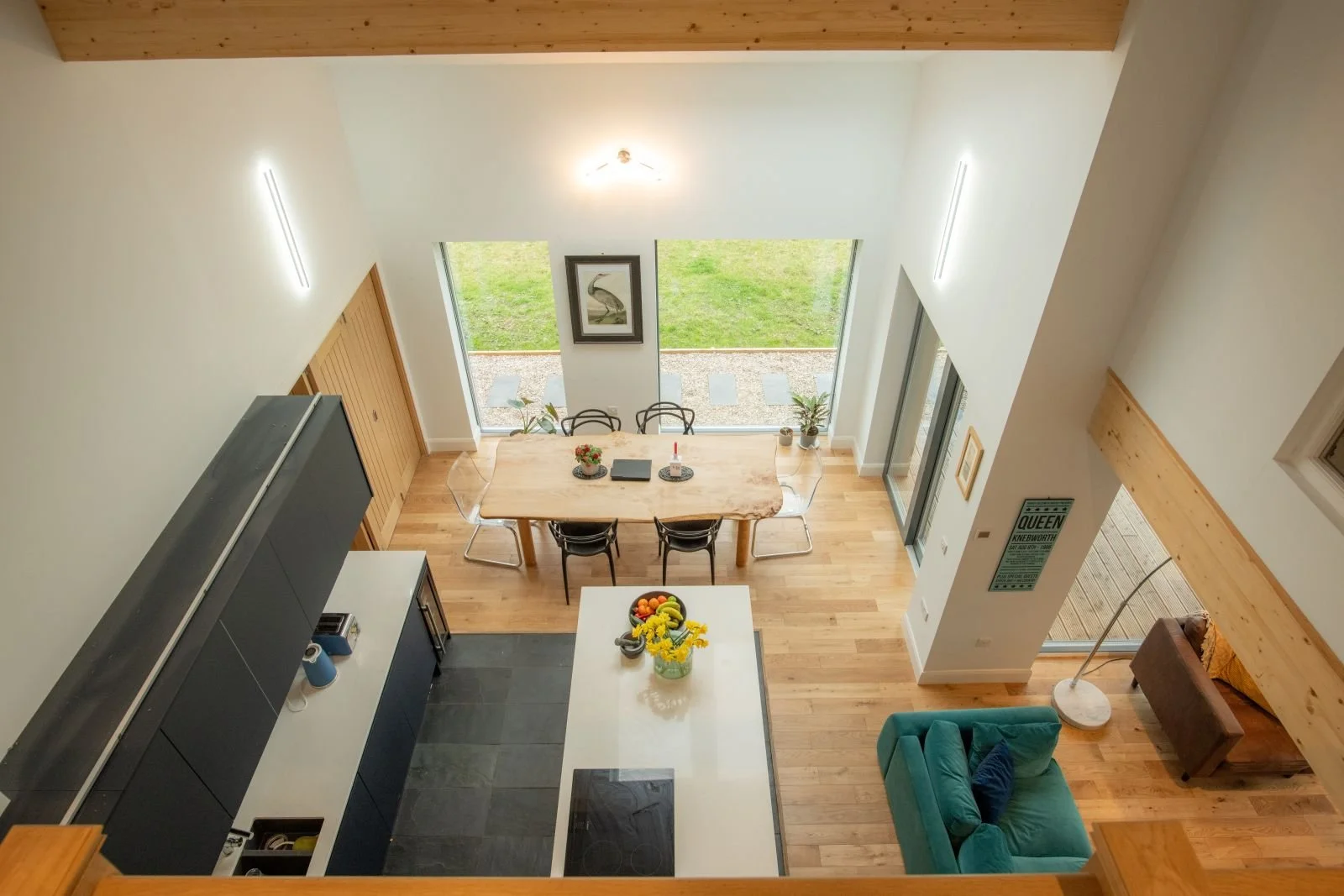 Modern dining and living area of a MAKAR home with a wooden dining table, black chairs, a kitchen island with flowers, a teal sofa, and large windows showing a garden outside.