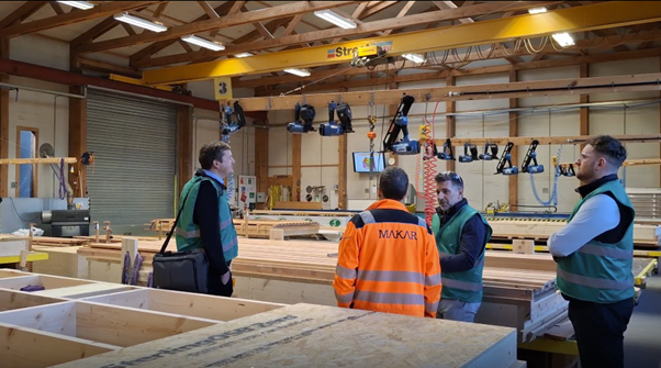 Four men in MAKAR's precision manufcature workshop, three wearing safety vests and one in an orange high-visibility jacket, engaged in conversation surrounded by panel creation for homebuilding projects.