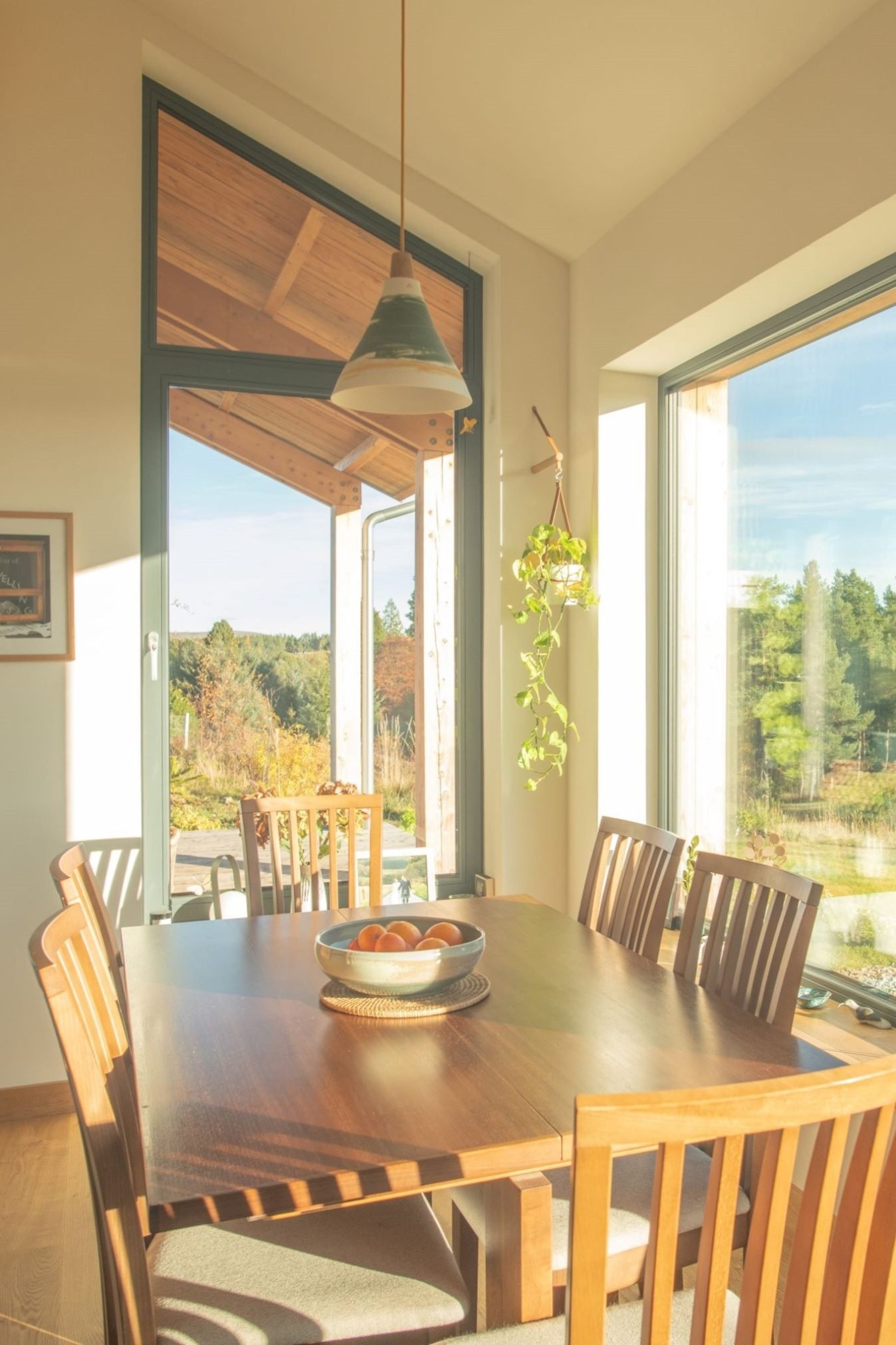 Sunlit dining room with wooden table, six chairs, a bowl of oranges, large windows, and a view of trees and sky outside.