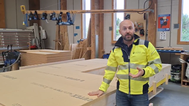 Man in a yellow safety jacket standing in a woodworking shop with large closed construction panels, part way through manufacture, and tools hanging on the wall.