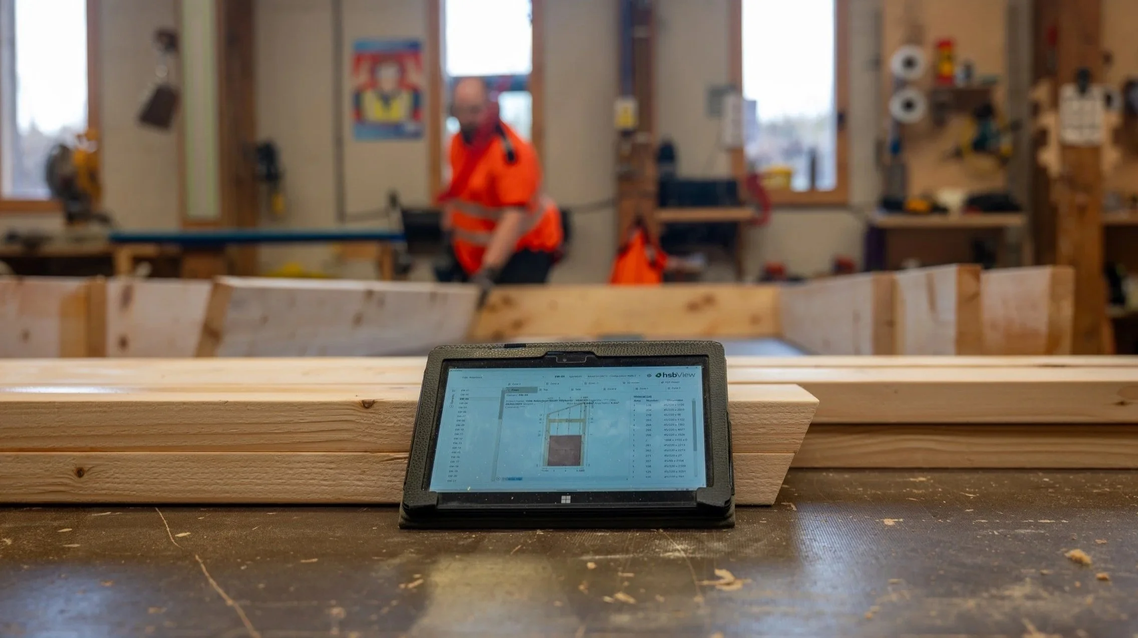 Close-up of a tablet showing hsbcad software for precision-manufacturing and live updates. The tablet is on a woodworking workbench displaying a construction plan, with a blurred woodworker in safety gear working in MAKAR's workshop background.