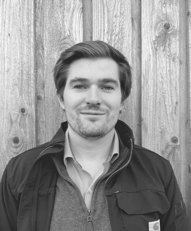 Young man with wavy hair smiling, wearing a jacket over a shirt and sweater, standing in front of a wooden wall.