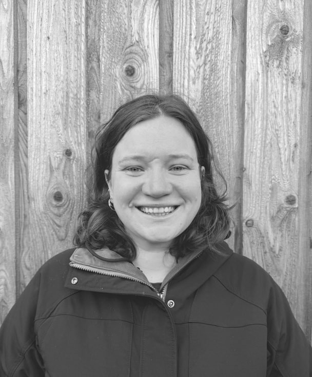 A woman with shoulder-length hair smiling, wearing a dark jacket, standing in front of a wooden fence.