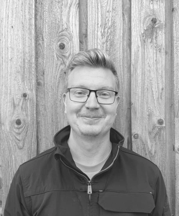 A young man with short, styled hair and glasses, smiling in front of a wooden fence.