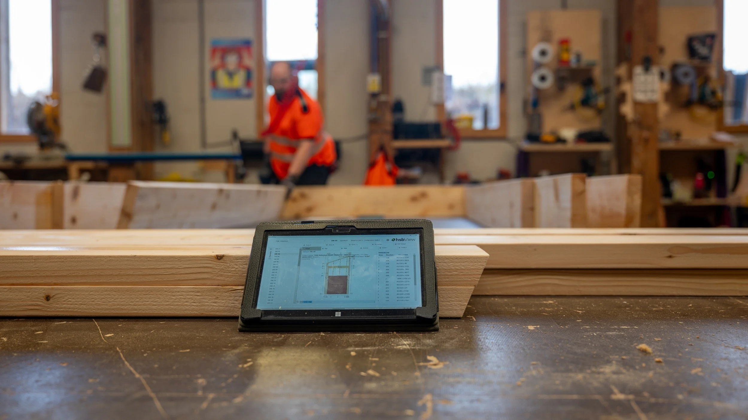 MAKAR's precision-manufacturing workshop with a tablet displaying a digital plan in the foreground and a person in an orange safety vest working in the background.