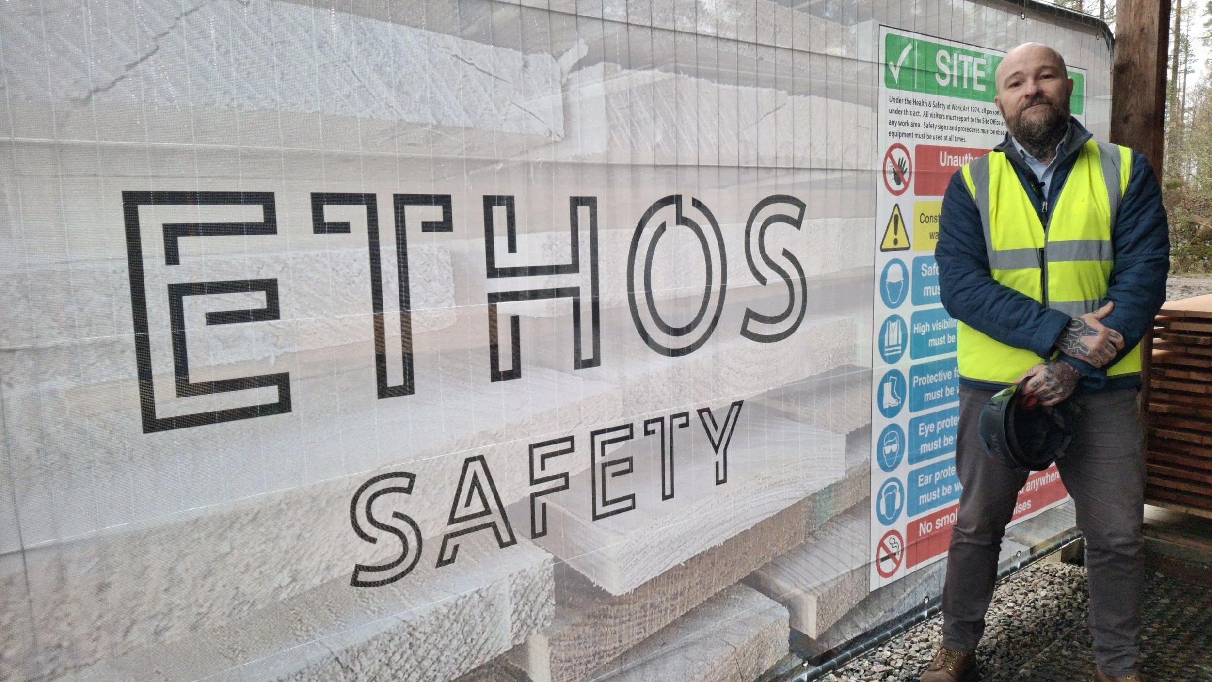 A man with tattoos in a yellow safety vest standing next to an Ethos Safety sign at a construction site.