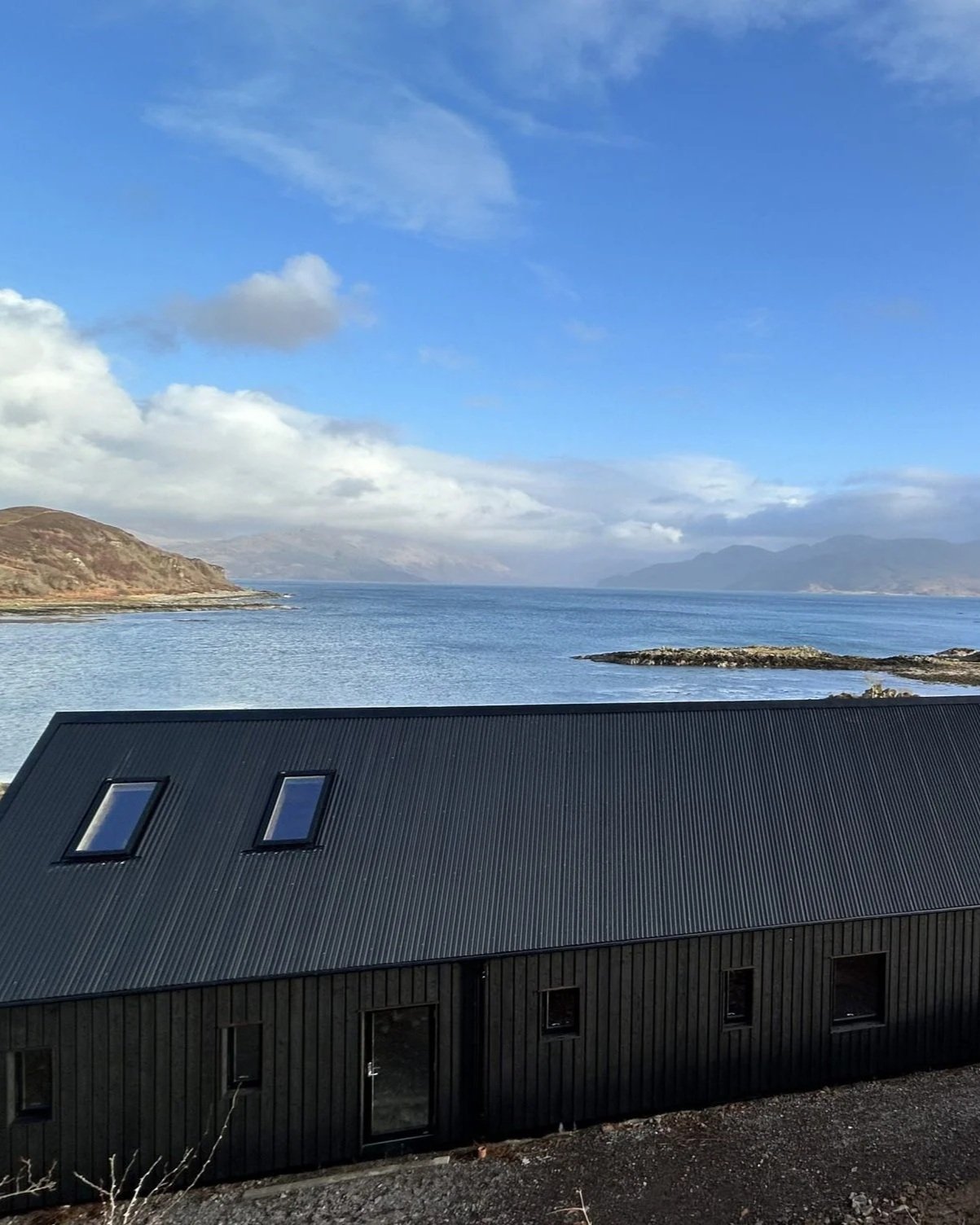 Black house with a metal roof and skylights overlooking a body of water with hills and mountains in the background under a partly cloudy sky.